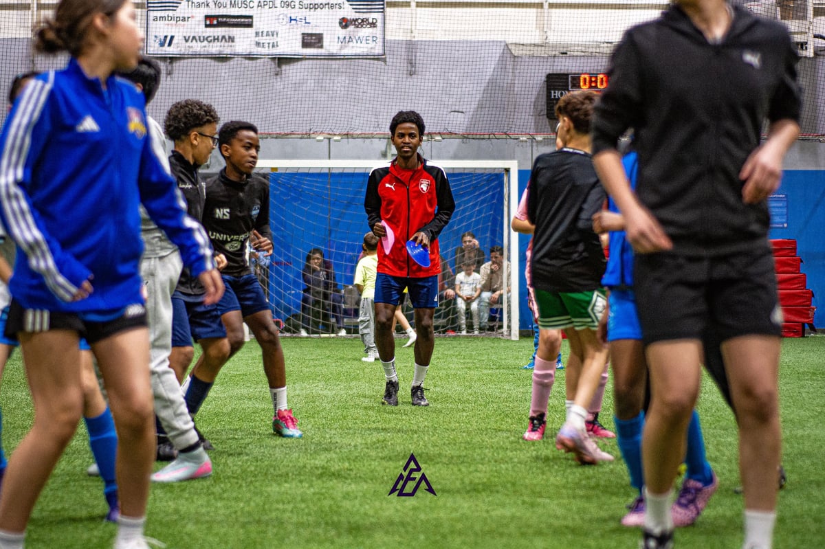 Youth soccer players in blue, black, and red jerseys playing on an indoor grass field with a blue soccer goal in the background