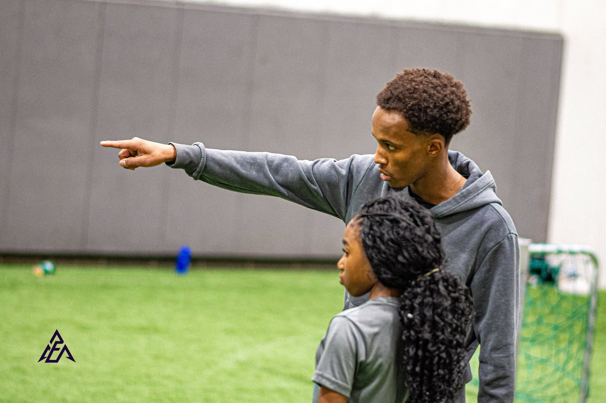 Adult pointing while instructing young child on indoor grass training field