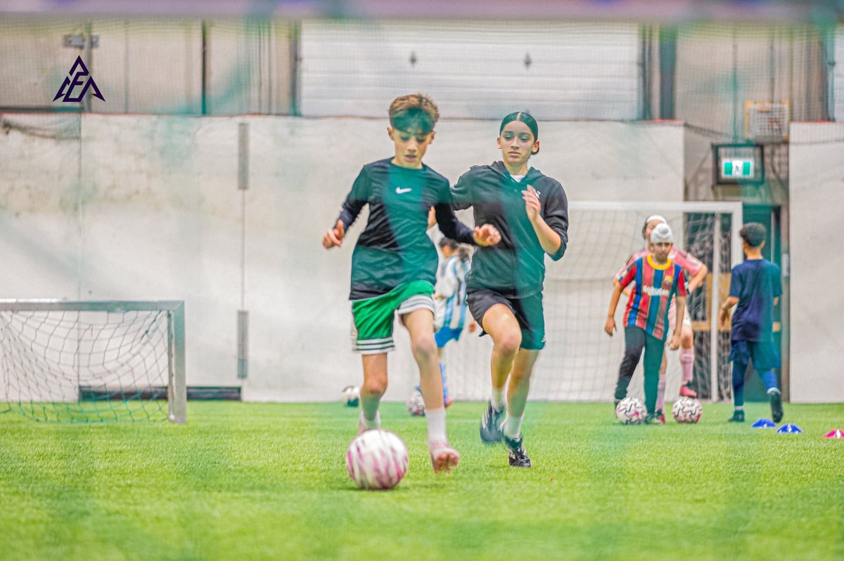 Two young soccer players in teal uniforms running with a ball during a youth sports match indoors