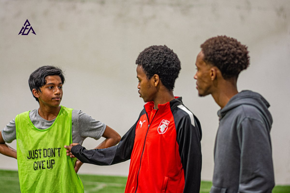 Three young men talking outdoors, one wearing a lime green shirt that says Just Dont Give Up, another in a red and black jacket, and a third in a gray hoodie