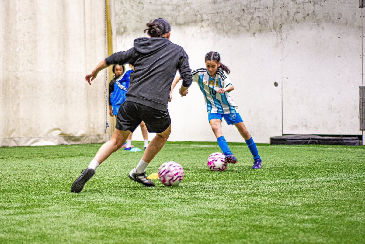 Adult soccer coach dribbling ball while young player in blue jersey watches during training on indoor grass field