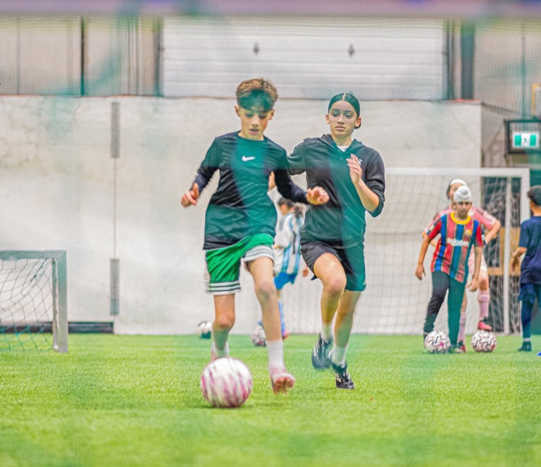 Two young soccer players in teal jerseys running after a ball during an indoor match with teammates and goals visible in background